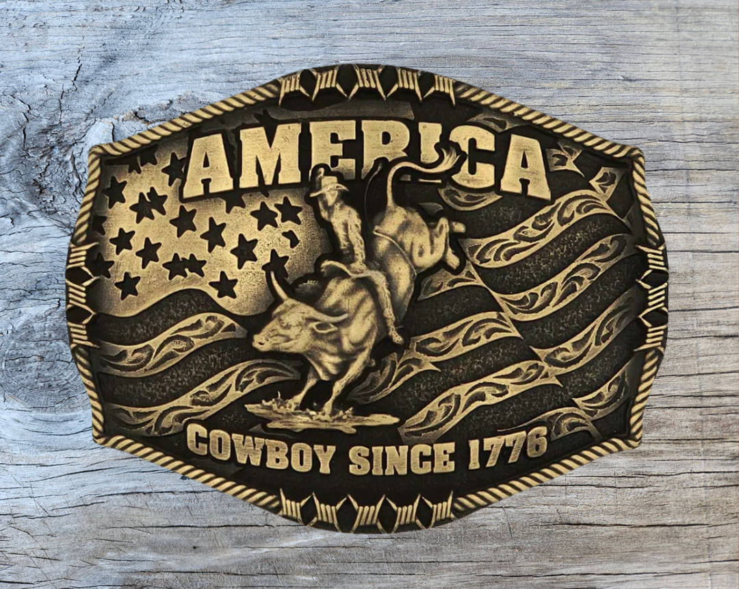 Stand for liberty and Western heritage for all with the Cowboy Since 1776 Attitude Buckle. The waving American flag provides the backdrop for the design. The word "America" sits above the bull rider figure. The words "Cowboy Since 1776" is beneath the bull rider figure. A roped border with barbed wire design creates the edge of the buckle, giving the piece a rustic look. Fits 1.5 inch belts. Find it online or at our shop just outside Nashville in Smyrna, TN.