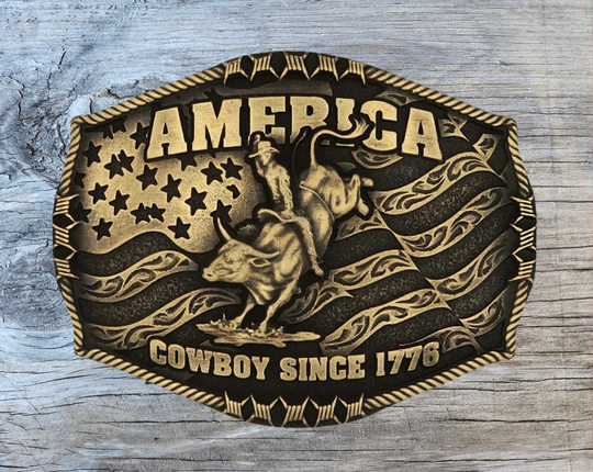Stand for liberty and Western heritage for all with the Cowboy Since 1776 Attitude Buckle. The waving American flag provides the backdrop for the design. The word "America" sits above the bull rider figure. The words "Cowboy Since 1776" is beneath the bull rider figure. A roped border with barbed wire design creates the edge of the buckle, giving the piece a rustic look. Fits 1.5 inch belts. Find it online or at our shop just outside Nashville in Smyrna, TN.