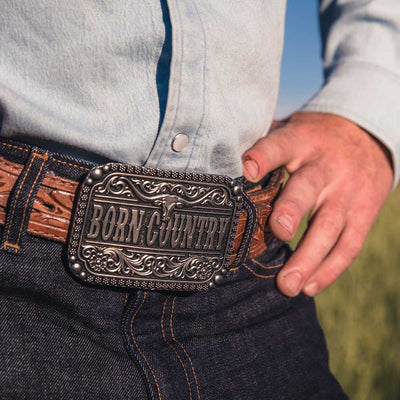 Wild, free, and born country. Show off your roots with this Silver-toned "Born Country" Buckle. With Born Country at the center of the buckle with a longhorn steer sitting above. Classic filigree highlights the top and bottom and additional silver-toned borders add texture. Flowers, and silver-toned beads border the outer edge. Fits 1 1/2" belts and is approx. 3" tall x 4" across. Available at our shop just outside Nashville in Smyrna, TN. Made by Montana Silversmith.