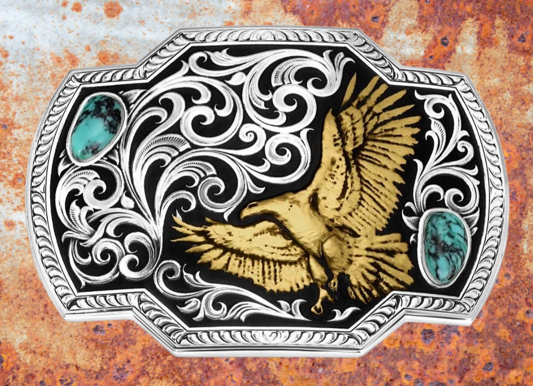 Take flight with the Tail Wind belt buckle! Featuring a black backdrop, a golden eagle soars through detailed silver filigree that mimics swirling winds. Crafted Turquoise stones add a pop of color in opposite corners of this rope-edged, rectangle buckle. Measuring about 4.25" wide by 3.25" tall, it fits belts up to 1.5" wide. Snag one at our Smyrna, TN shop—just a quick drive from downtown Nashville! Made by Montana Silversmith.