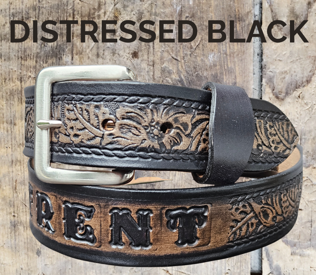 A western-style leather belt in a distressed black finish, featuring tooled floral detailing embossed on the strap, shown on a rustic wood background with text overlays.