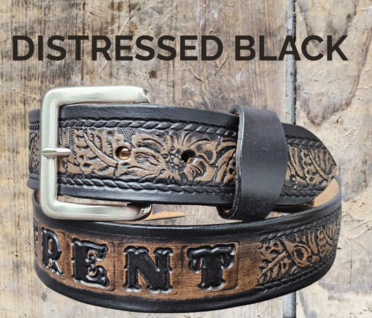 A western-style leather belt in a distressed black finish, featuring tooled floral detailing embossed on the strap, shown on a rustic wood background with text overlays.