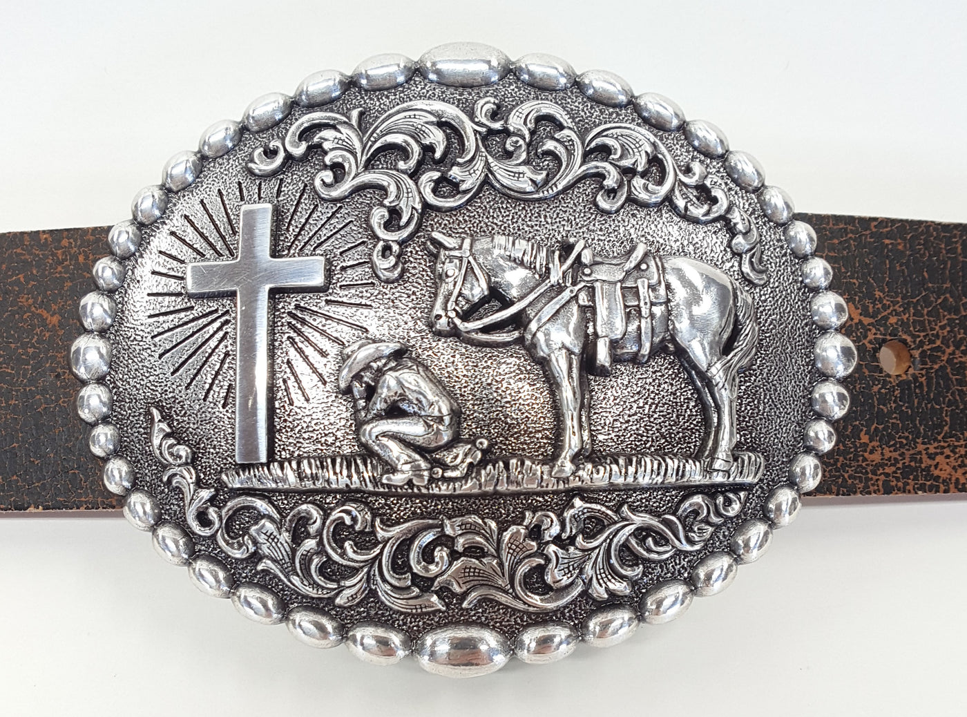 Nocona buckle with beaded edge. Cowboy kneeling at Cross with his trusted horse behind him. A raised floral design perfectly complements this reverent design. Measures 3-1/8" tall by 4" wide  Available online and in our shop in Smyrna, TN, just outside of Nashville